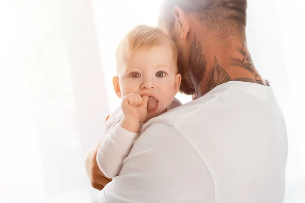 Vista posteriore del giovane uomo tatuato che tiene il bambino guardando la fotocamera con la mano in bocca — Foto stock