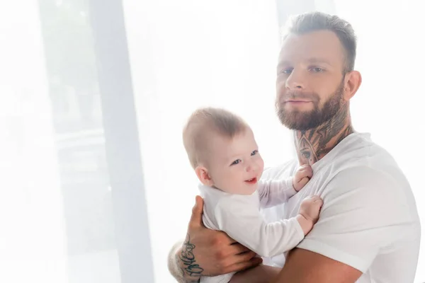 Joven hombre tatuado mirando a la cámara mientras sostiene alegre bebé hijo - foto de stock