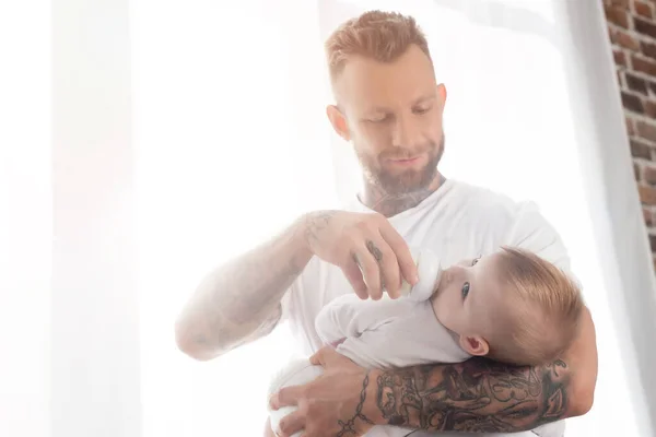 Joven barbudo padre alimentación bebé niño mientras de pie cerca de ventana - foto de stock