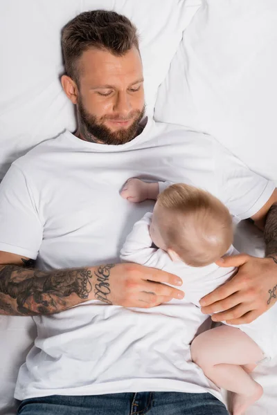 Vue de dessus du jeune homme tatoué et fils de bébé dans la barboteuse de bébé couché dans le lit — Photo de stock