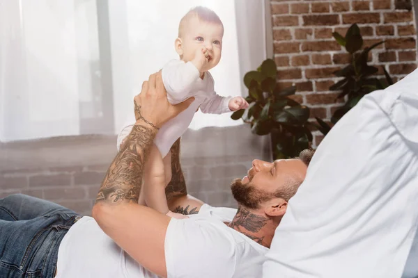 Enfoque selectivo de joven hombre tatuado sosteniendo excitado bebé niño mientras está acostado en la cama - foto de stock