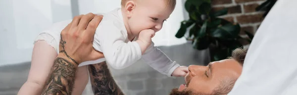 Orientación panorámica de la mentira hombre tatuado sosteniendo hijo bebé en el dormitorio - foto de stock