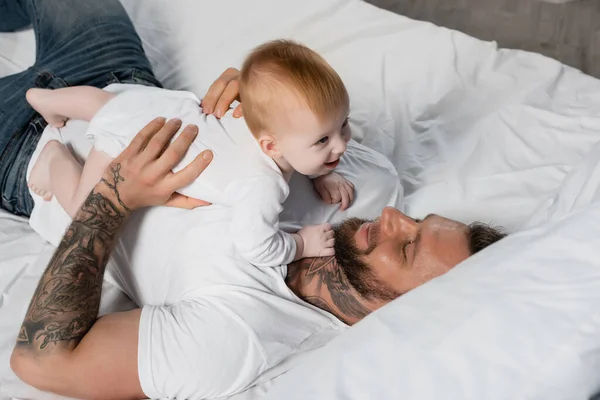 Vista aérea del joven hombre tatuado divirtiéndose con el bebé mientras está acostado en la cama - foto de stock