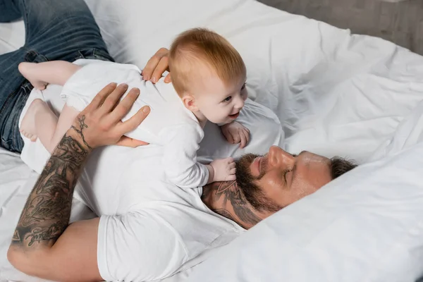 Visão aérea do jovem homem tatuado se divertindo com o bebê enquanto deitado na cama — Fotografia de Stock