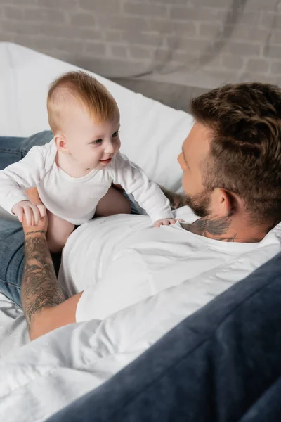 Vue aérienne d'un jeune homme tatoué tenant un enfant en bas âge assis au lit — Photo de stock