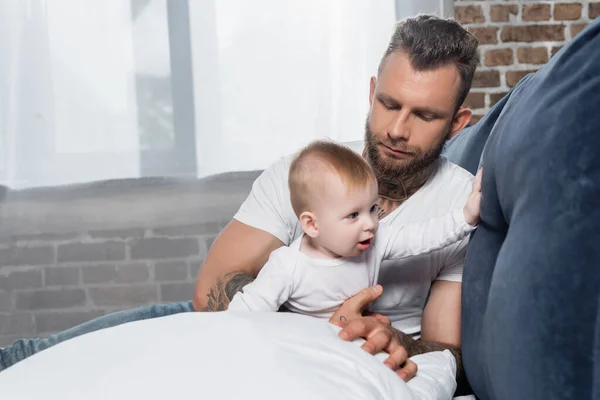 Selective focus of young bearded man sitting on bed near infant child — Stock Photo