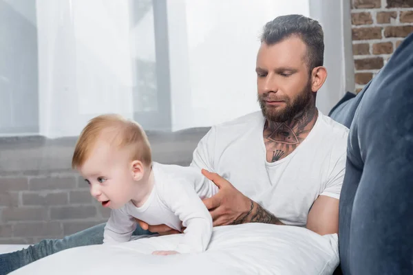 Joven barbudo hombre tocando infante hijo arrastrándose en cama en bebé mameluco - foto de stock