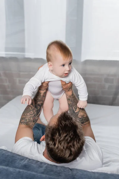 Selective focus of tattooed man holding baby boy while sitting in bed — Stock Photo