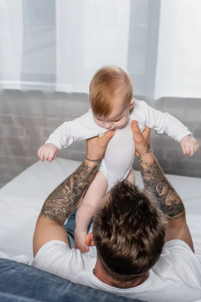 Vista aérea del hombre tatuado sosteniendo al bebé en un mameluco mientras está sentado en la cama - foto de stock