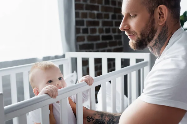 Barbudo, hombre tatuado que apoya al niño de pie en la cuna - foto de stock