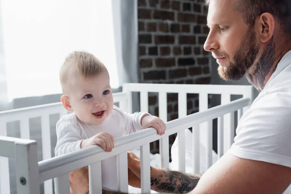 Junger bärtiger Mann stützt aufgeregten kleinen Jungen, der im Kinderbett steht — Stockfoto