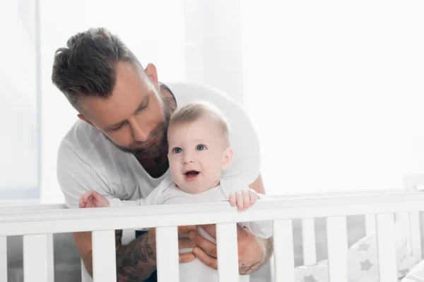 Jeune père soutenant enfant excité debout dans un lit bébé avec bouche ouverte — Photo de stock