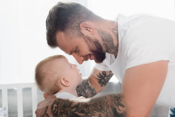 Vista lateral de alegre hombre tatuado y bebé niño mirándose cara a cara - foto de stock