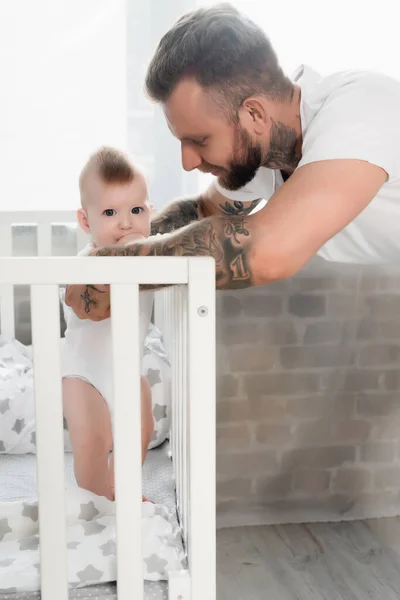 Homem tatuado apoiando criança infantil em pé no berço e de mãos dadas na boca — Fotografia de Stock
