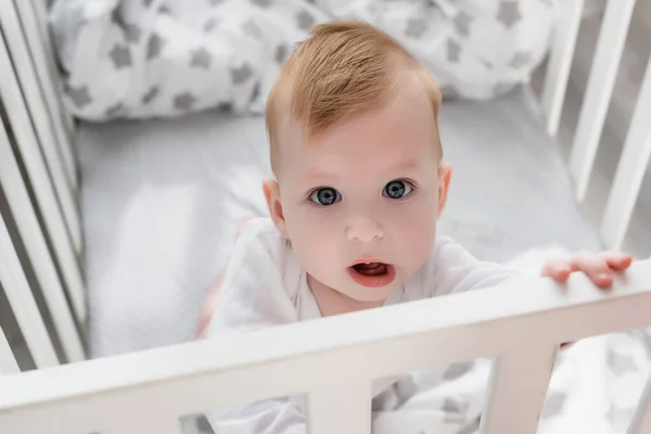 Overhead view of baby boy looking at camera while standing in crib with open mouth — Stock Photo