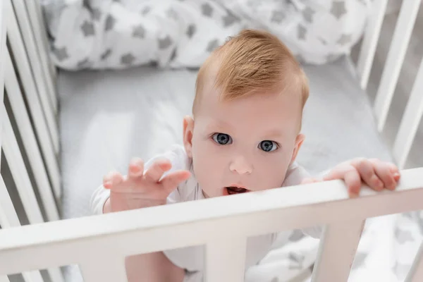Selective focus of infant boy looking at camera while standing in crib with outstretched hand — Stock Photo
