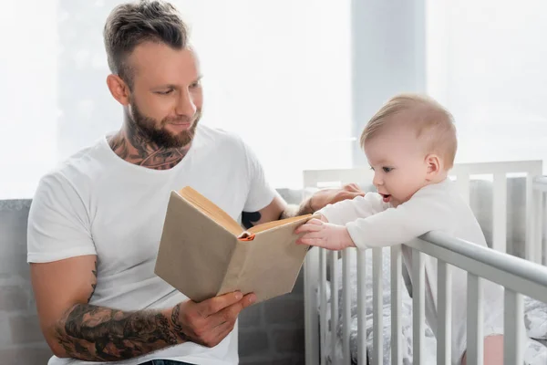 Infant boy standing in crib and touching book in hands of young father — Stock Photo