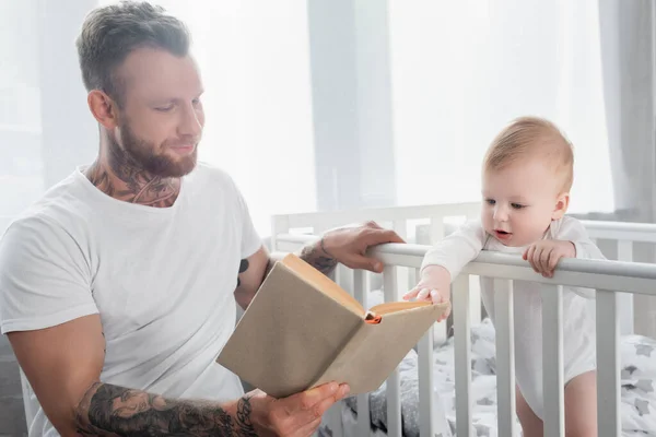 Молодий татуйований батько читає книгу дитині хлопчика, що стоїть у ліжку — стокове фото