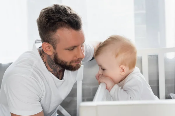 Молодой татуированный мужчина смотрит на младенца, стоящего в кроватке — стоковое фото