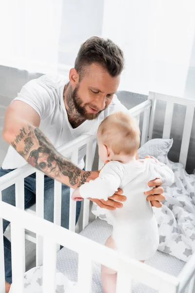 Selective focus of young tattooed man supporting infant son standing in crib — Stock Photo