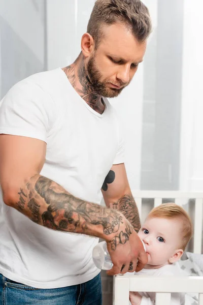 Joven tatuado hombre alimentación infantil niño de pie en cuna - foto de stock