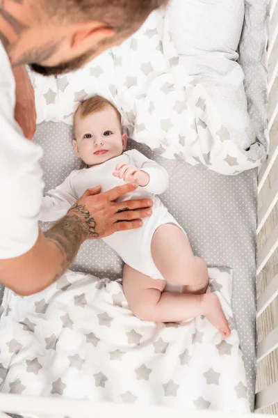 Vue aérienne du père touchant le fils de bébé couché dans la crèche dans la barboteuse de bébé — Photo de stock