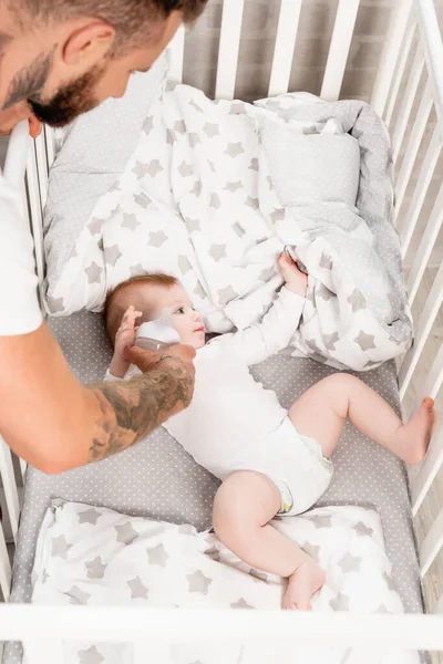 Vista de ángulo alto del hombre joven tatuado alimentando a un niño lactante acostado en la cuna - foto de stock