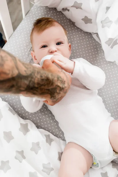 Selektiver Fokus tätowierter Vater füttert im Kinderbett liegenden Jungen im Babystrampler — Stockfoto