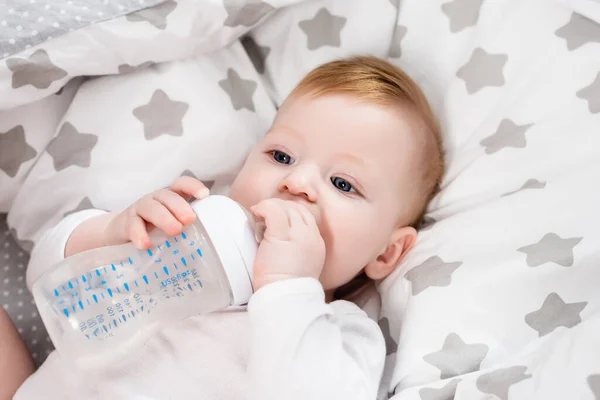 Infant boy holding baby bottle while lying on bedding with stars print — Stock Photo