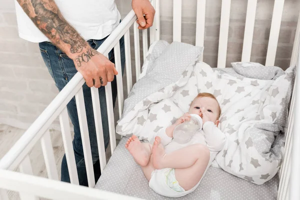 Hombre tatuado de pie cerca de su hijo pequeño acostado en una cuna con biberón - foto de stock