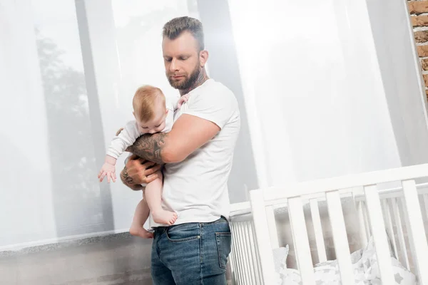 Татуйований чоловік тримає хлопчика, стоячи біля ліжечка у вікні — стокове фото