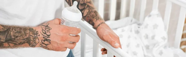 Partial view of tattooed man holding baby bottle while standing near crib, horizontal concept — Stock Photo
