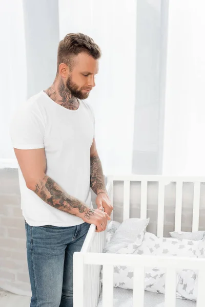 Jeune homme tatoué en t-shirt blanc debout près du lit à la maison — Photo de stock
