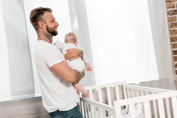 Young father holding infant boy while standing at crib near window — Stock Photo