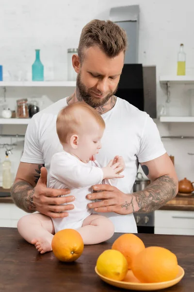 Jeune homme tatoué tenant bébé fils assis sur la table de cuisine près des oranges — Photo de stock