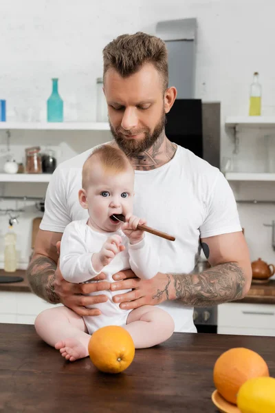 Young tattooed man near infant son holding spoon near mouth while sitting on kitchen table — Stock Photo