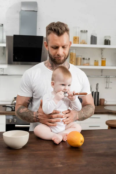 Niño pequeño sosteniendo cuchara cerca de la madre mientras está sentado en la mesa de la cocina cerca del padre joven - foto de stock