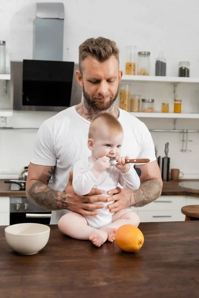 Infant child holding spoon near mother while sitting on kitchen table near young father — Stock Photo