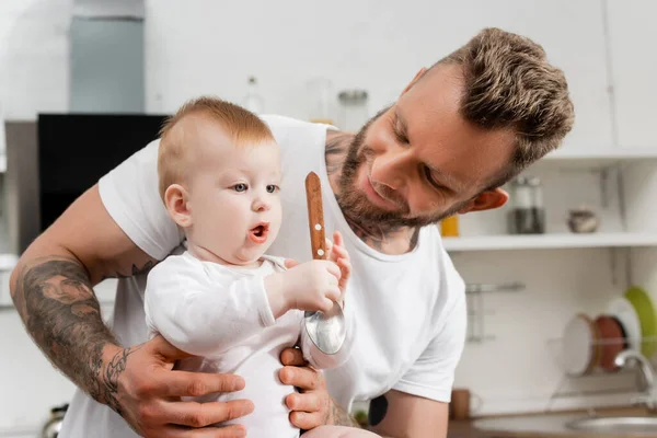 Infant boy holding spoon near young, bearded father in kitchen — Stock Photo