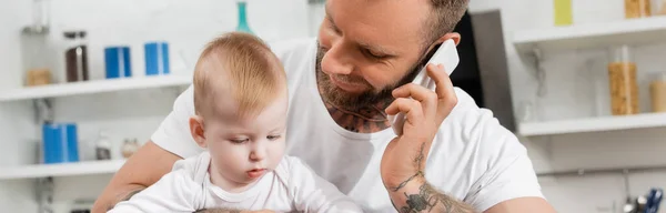 Panoramic orientation of young man talking on mobile phone near infant kid in kitchen — Stock Photo