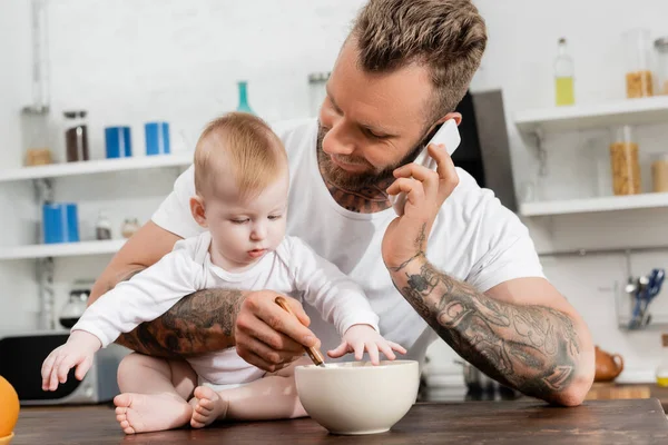 Joven padre hablando en el teléfono inteligente mientras se mezcla el desayuno en un tazón cerca de hijo pequeño - foto de stock