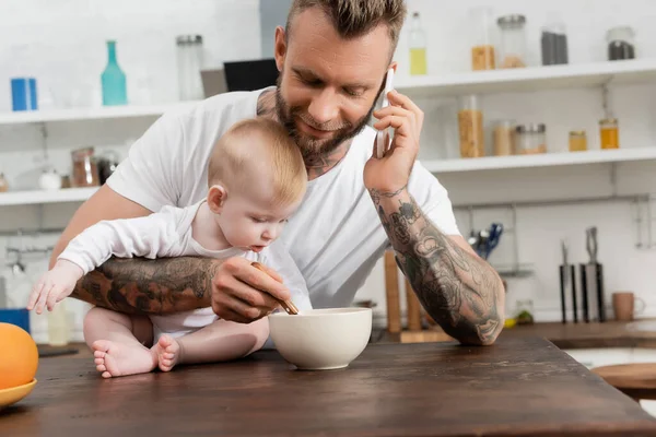 Joven barbudo hombre hablando en el teléfono inteligente cerca de niño y tazón con desayuno en la cocina - foto de stock