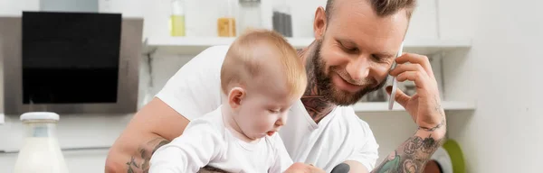 Horizontales Bild eines jungen Mannes, der während des Frühstücks mit seinem kleinen Sohn in der Küche mit dem Smartphone spricht — Stockfoto