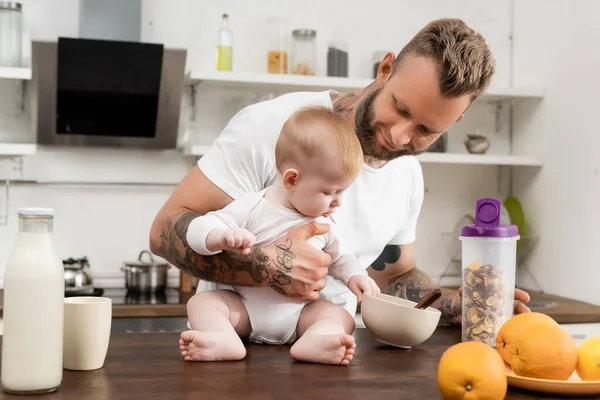 Bébé garçon toucher bol avec petit déjeuner tout en étant assis sur la table de cuisine près du jeune père tatoué — Photo de stock