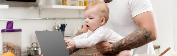 Panoramic shot of infant kid touching laptop near tattooed father in kitchen — Stock Photo