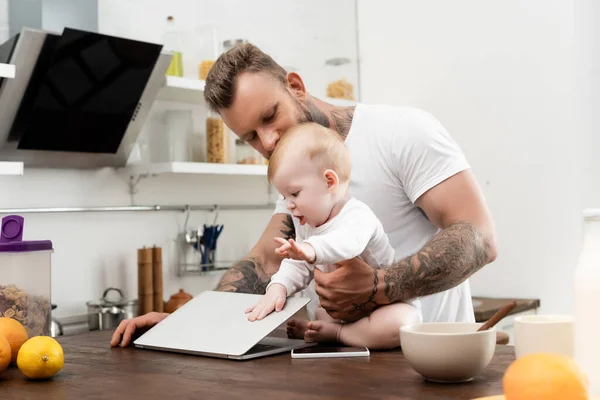 Menino bebê tocando laptop enquanto sentado na mesa da cozinha perto do pai tatuado — Fotografia de Stock