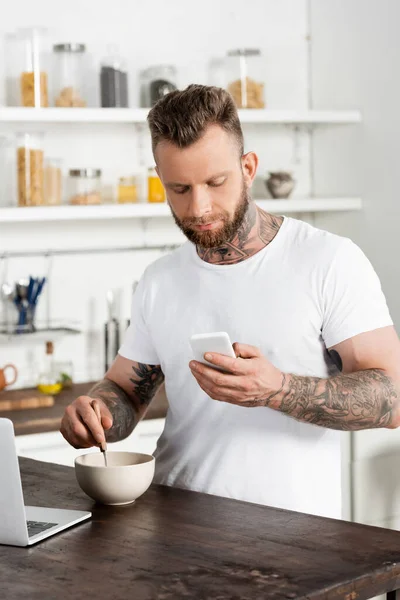 Bearded, tattooed freelancer mixing breakfast while chatting on mobile phone in kitchen — Stock Photo