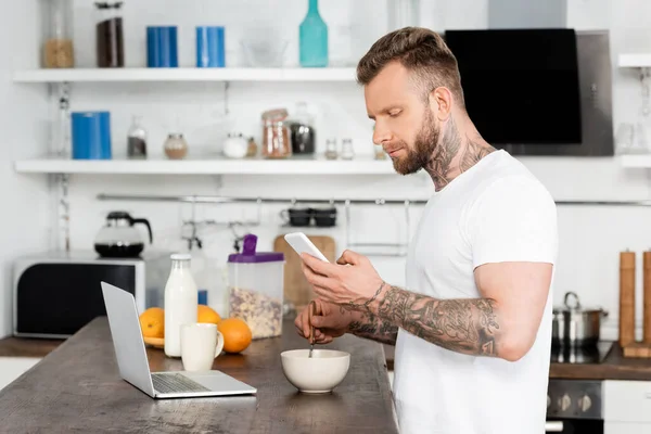 Tattooed freelancer chatting on smartphone near laptop during breakfast in kitchen — Stock Photo
