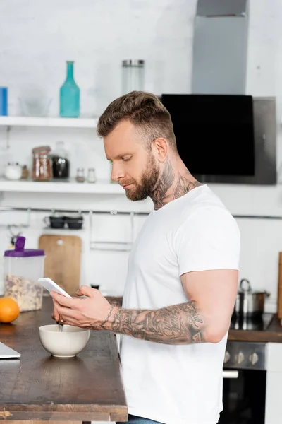 Молодой татуированный мужчина в белой футболке болтает на смартфоне во время завтрака на кухне — стоковое фото