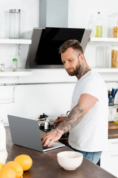 Young, bearded freelancer in white t-shirt using laptop near oranges and bowl in kitchen — Stock Photo